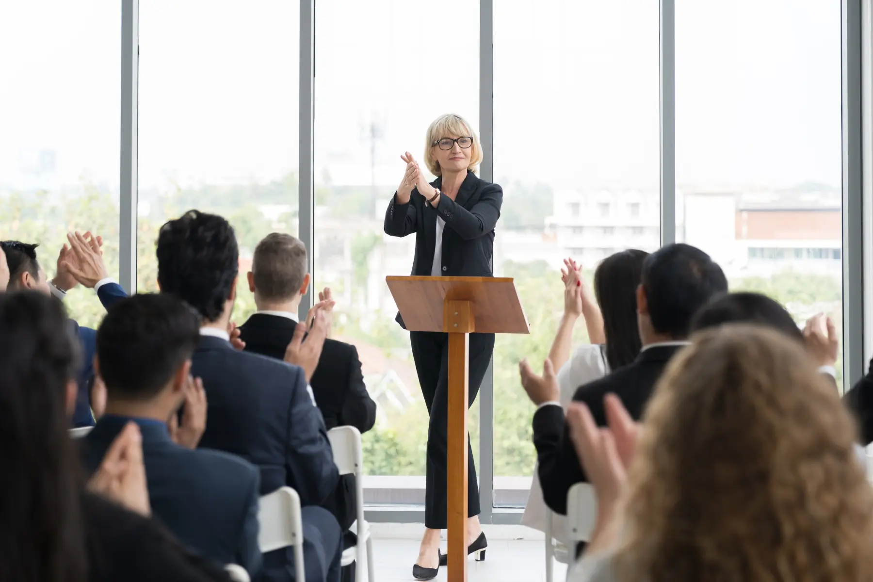 GettyImages-1414351216 A person at a podium claps while an audience seated indoors also applauds. Large windows in the background show an urban landscape.