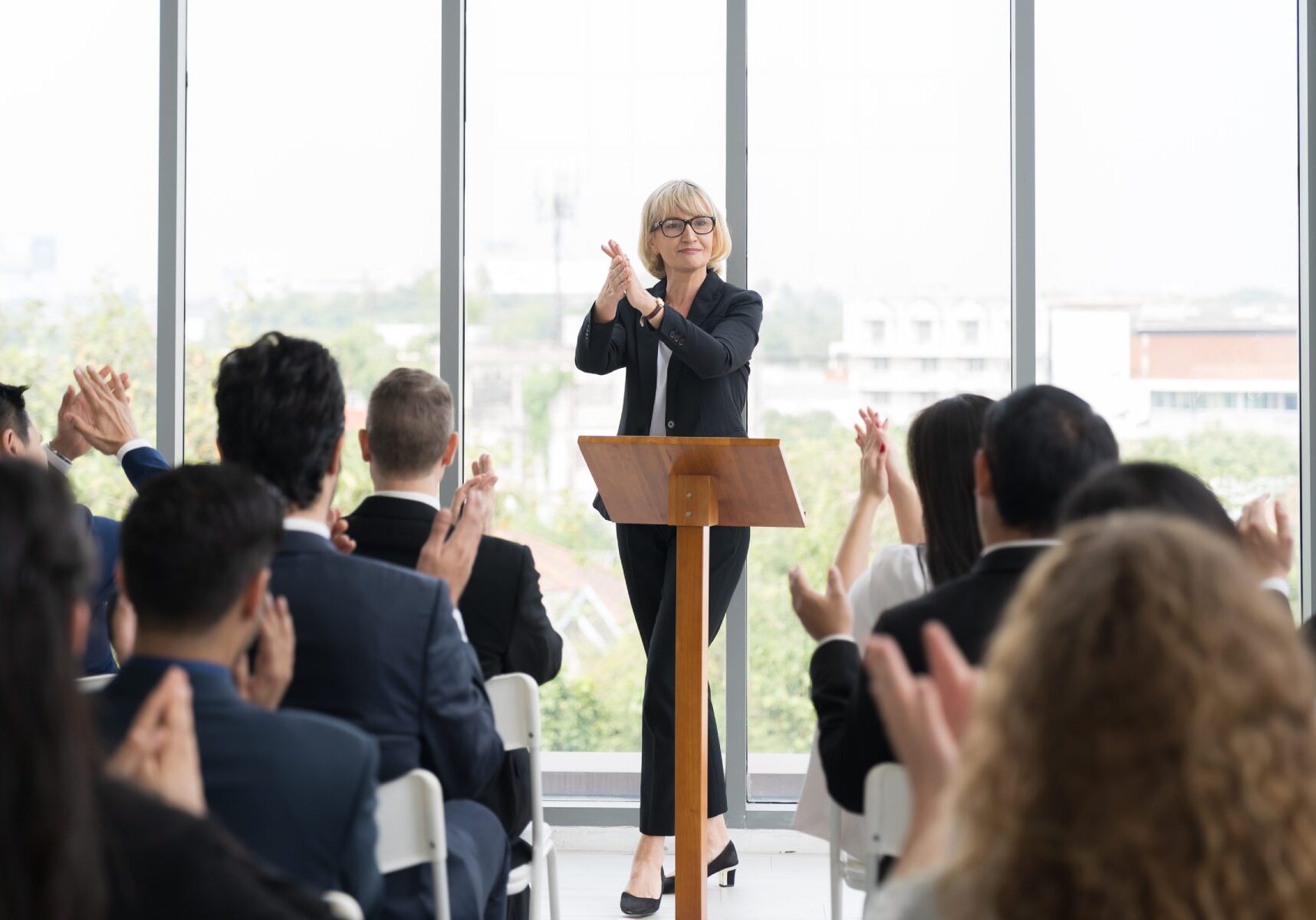 A person at a podium claps while an audience seated indoors also applauds. Large windows in the background show an urban landscape.